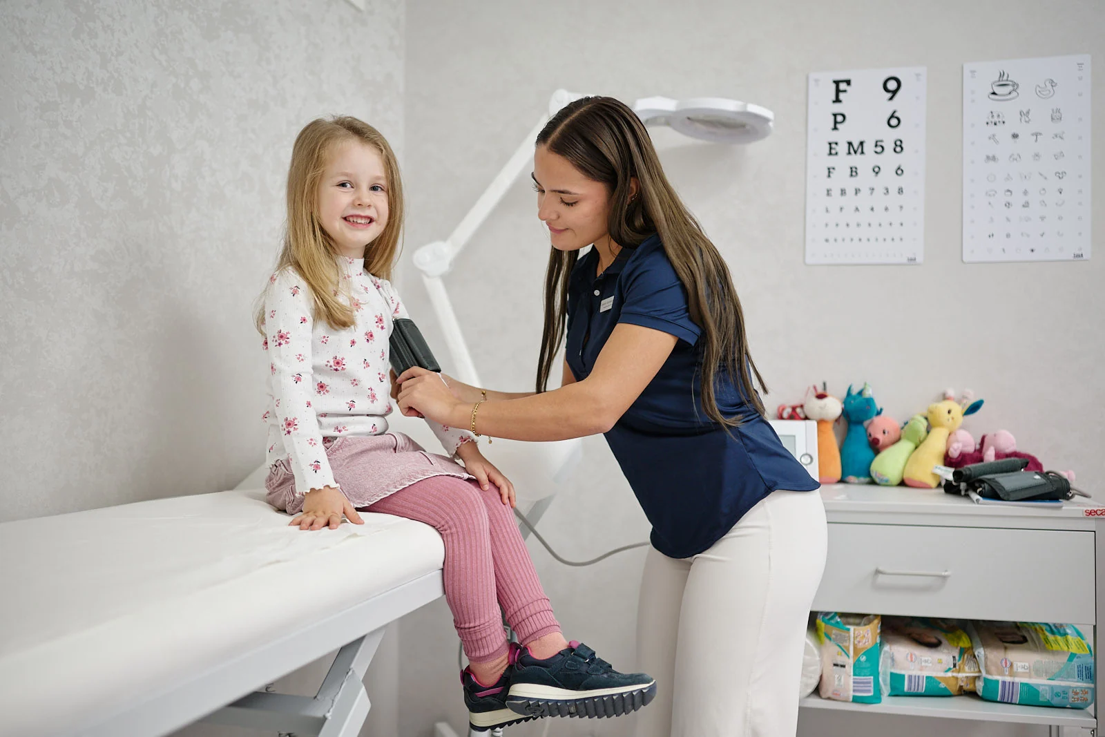 Praxisassistentin misst einem lachenden Mädchen den Blutdruck im Behandlungszimmer der Kinderarztpraxis Chur West